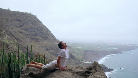 At-the-precipice-of-a-cliff,-the-man-practices-a-dog-pose,-facing-the-ocean,-taking-in-the-sea-breeze-during-his-yoga-journey-across-the-islands